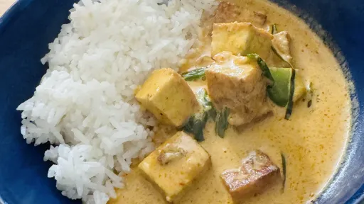 Steaming yellow curry with tofu and greens next to white rice in a blue bowl.