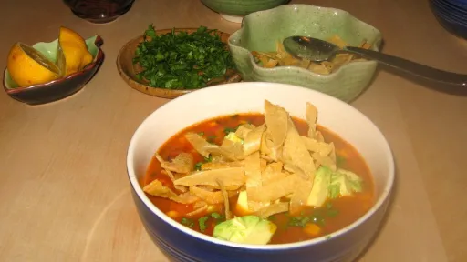 Blue bowl of tortilla soup with avocado, tortilla strips, chopped herbs, and lemon wedges.