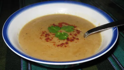 Creamy light brown lentil soup with a spoon, garnished with red spice and a green herb in a blue-rimmed bowl.