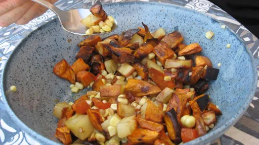 Close-up of a hand scooping roasted sweet potato and corn hash from a blue bowl.