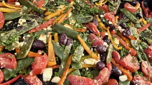 A smiling woman wearing glasses and an apron holds a large bowl of fresh salad in a bright, modern kitchen.