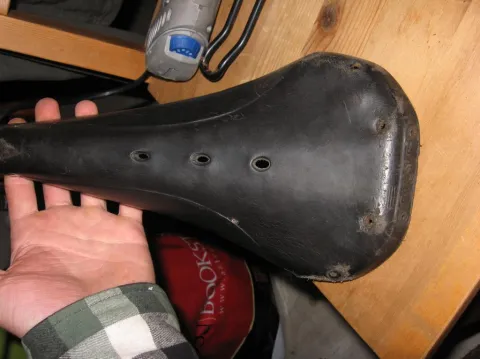 Hand holding a worn black bicycle saddle, leather only.