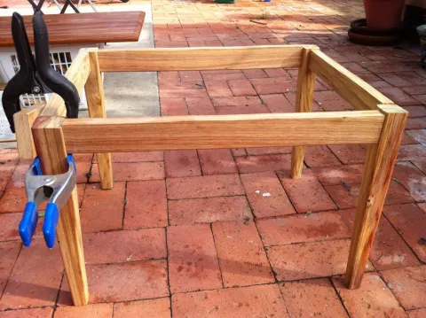 Wooden table frame being assembled with a clamp on a brick patio.