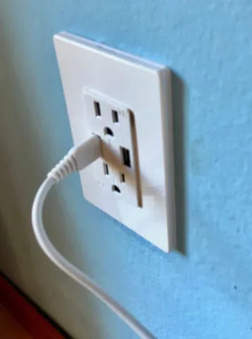 White wall outlet on a blue wall, with a white charging cable plugged into one of the USB socket.