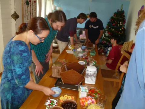 Group of people decorating gingerbread houses with colorful candies. Christmas tree in background.