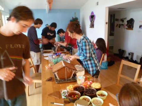 Group of people decorating gingerbread houses at a long table with various candies.