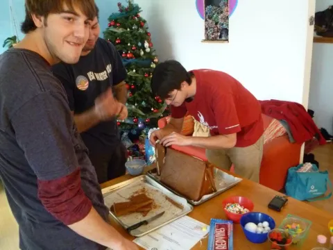Three men decorating a gingerbread house on a table, a Christmas tree in the background.